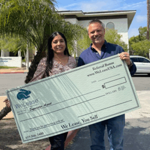 Billy Colestock and Yesenia Nogales standing outside the WeLease Property Management office in San Diego, holding a large check representing the WeLease, You Sell Realtor Referral Program.
